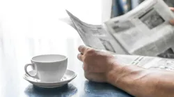 person reading newspaper near window while drinking a hot beverage