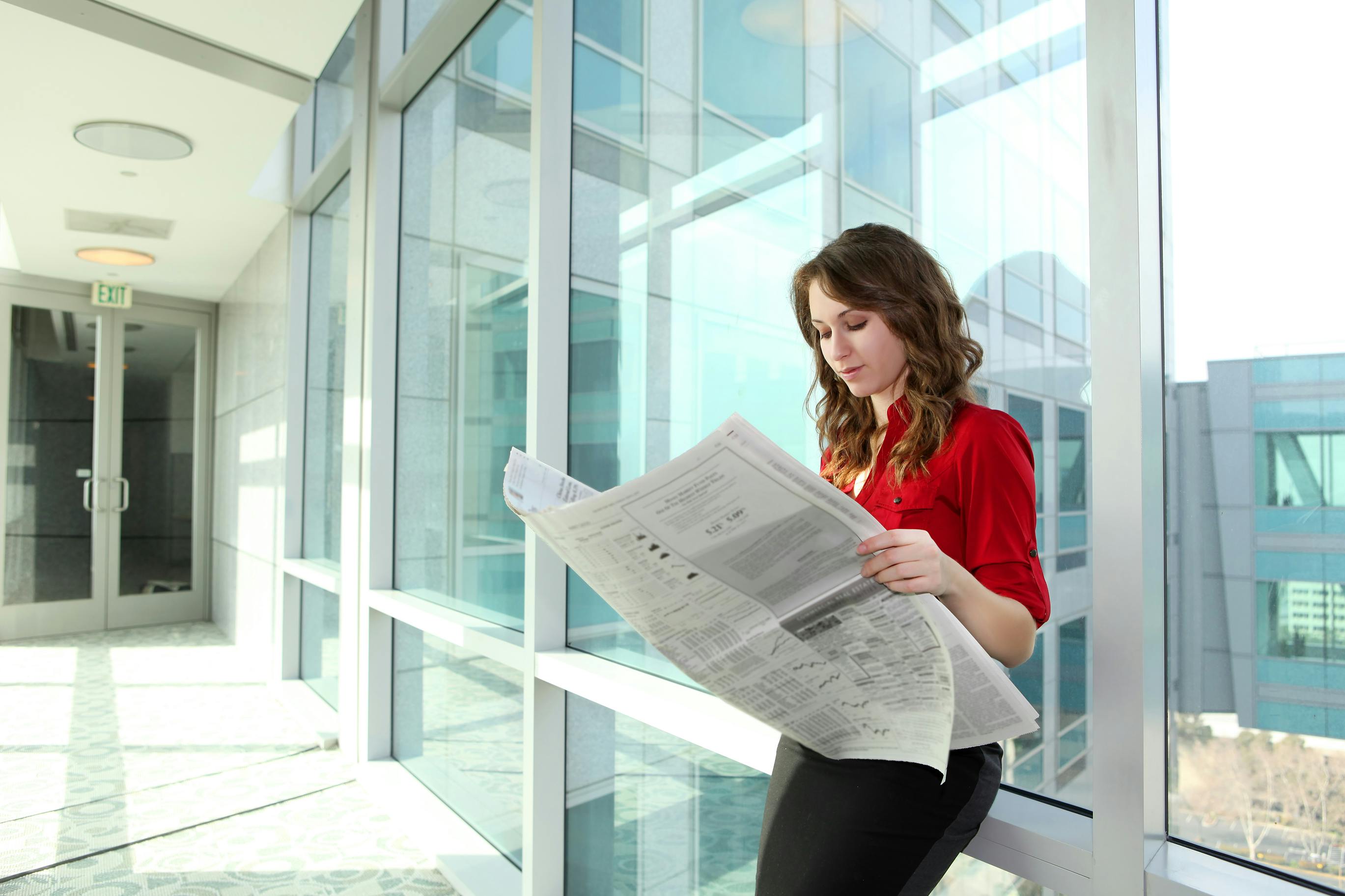 woman in professional attire reading a newspaper while leaning against a wall of window