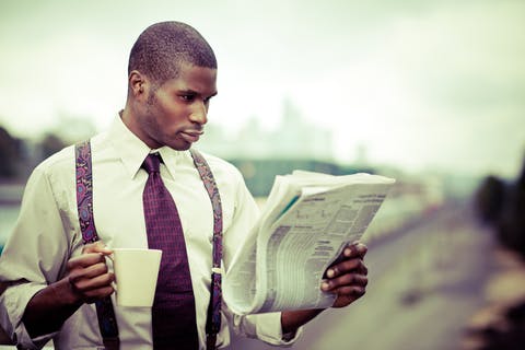 man in a collared shirt and suspenders reading the newspaper and holding a mug