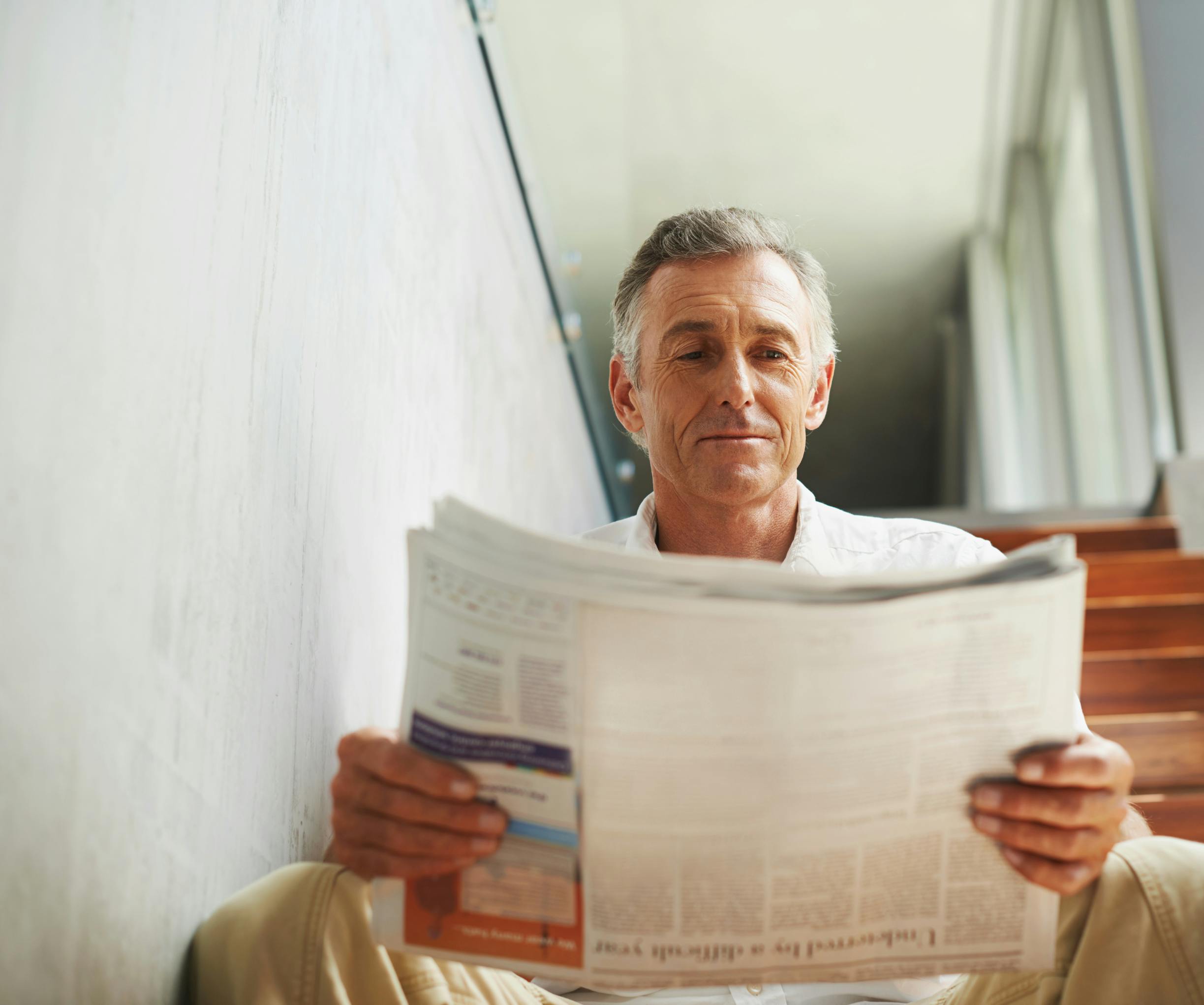 older white man sitting on wooden steps indoors reading a newspaper