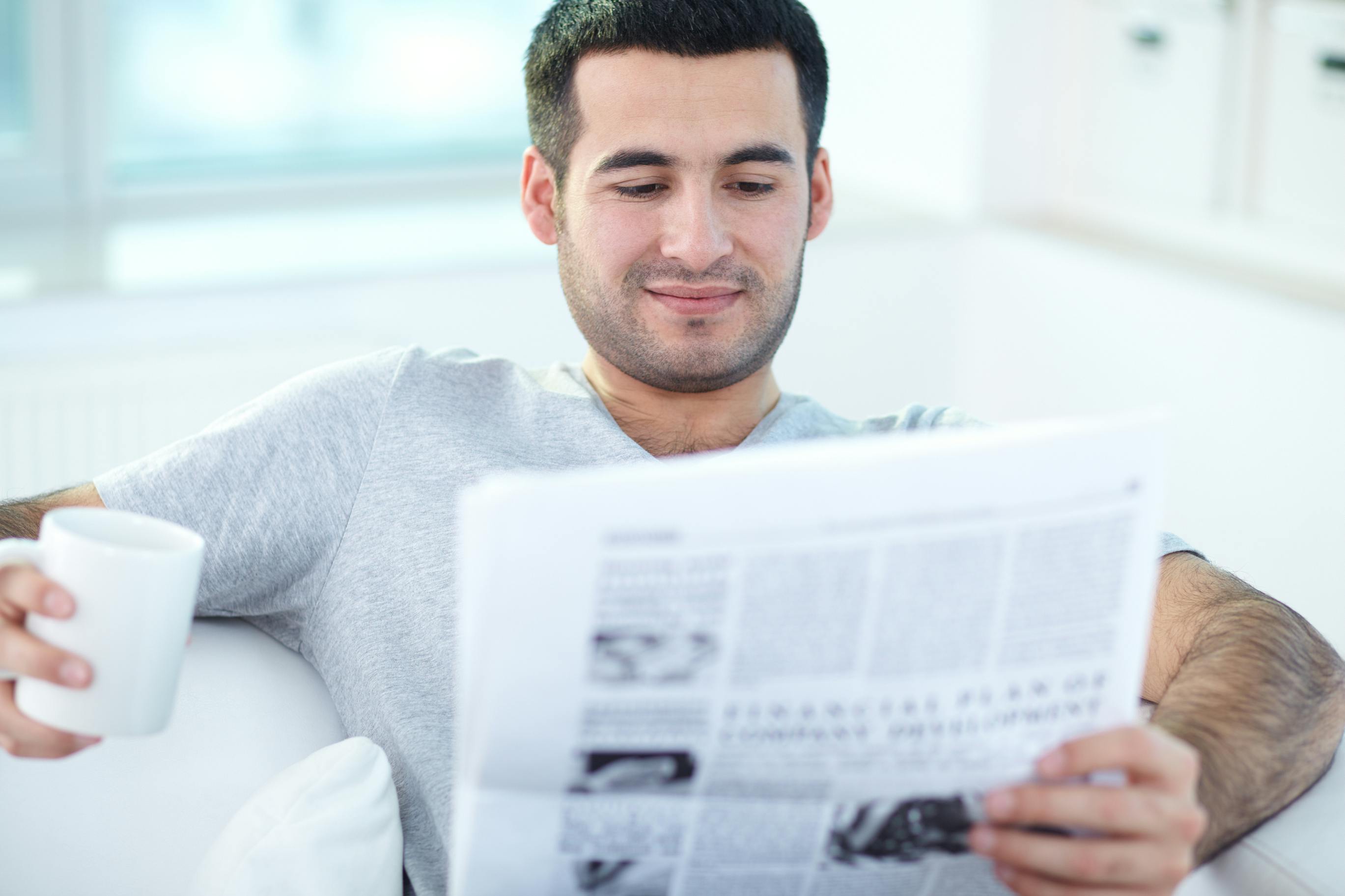 younger man reading newspaper on couch holding coffee mug