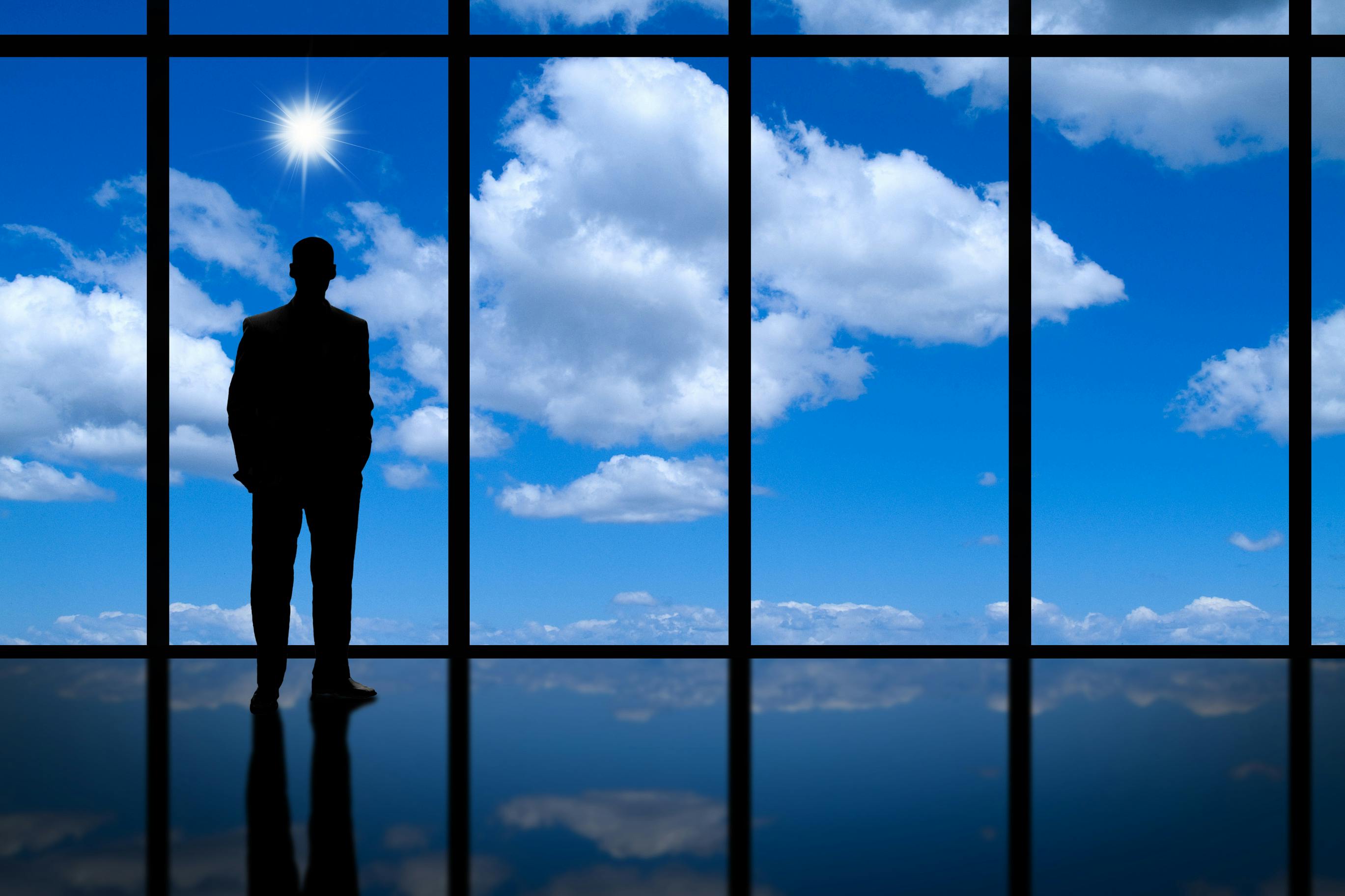 businessman silhouette looking through floor to ceiling windows of clouds and a bright blue sky