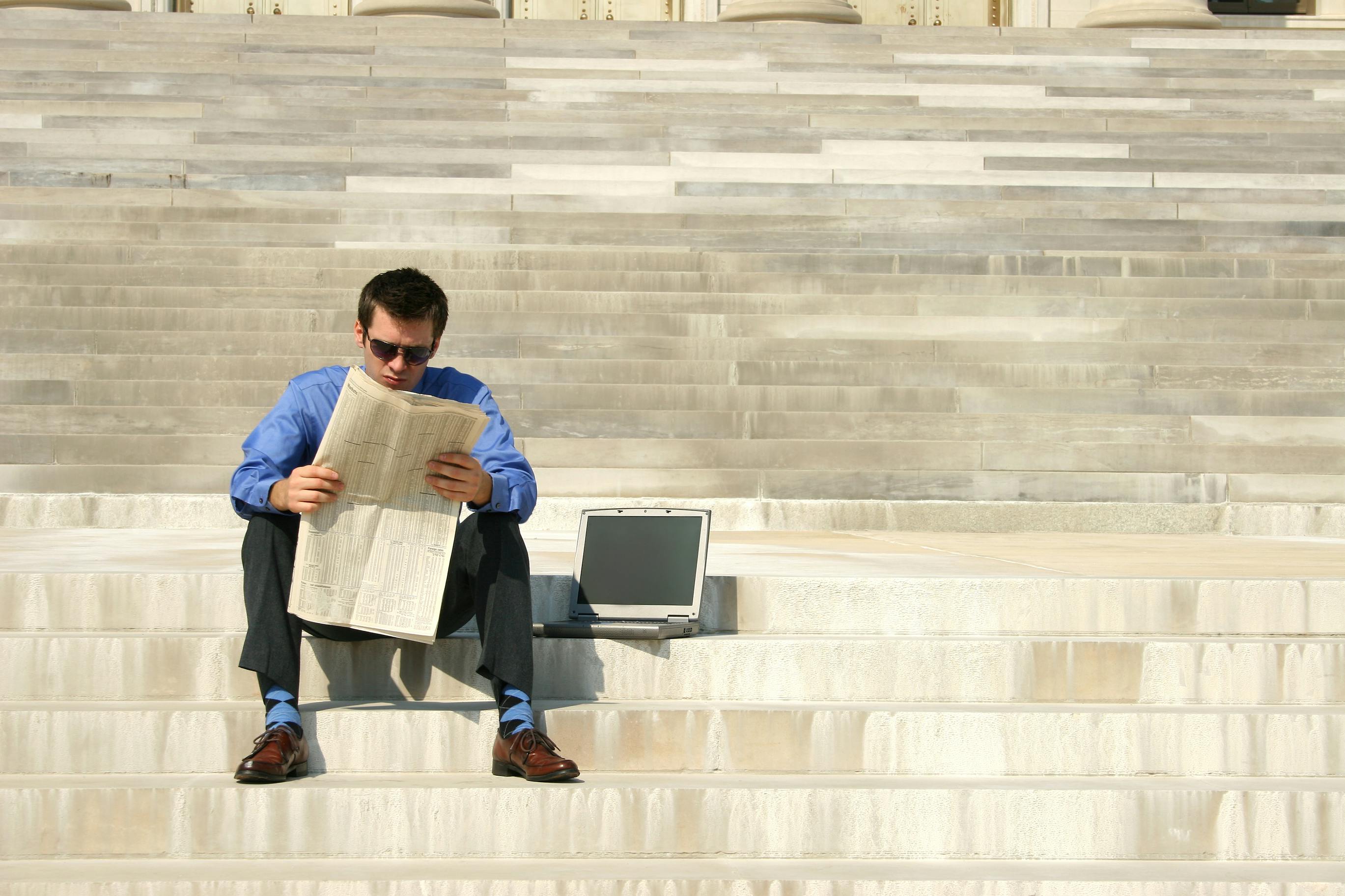 man in business attire sitting outside on the steps reading a newspaper with a laptop next to him