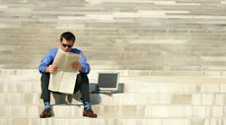 man in business attire sitting outside on the steps reading a newspaper with a laptop next to him