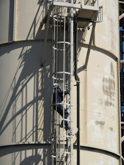 Photo of a worker climbing a caged ladder on the exterior of a metal structure Photo of a worker climbing a caged ladder on the exterior of a metal structure