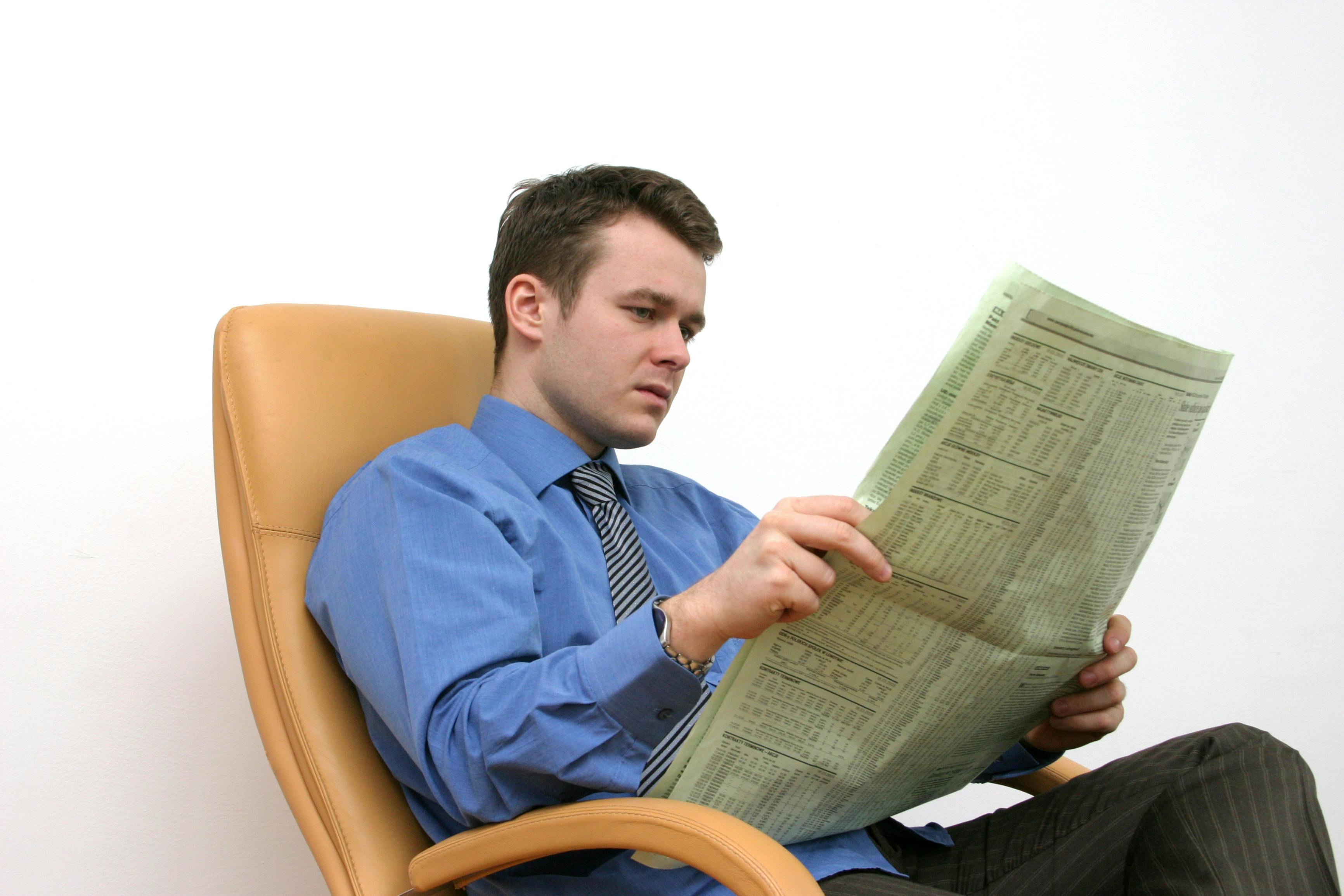 young man in a shirt, tie and dress slacks reading a newspaper in a beige leather desk chair