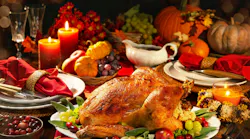 Picture of a tablescape with a turkey on a platter and formal place settings, gravy boat, candles, wine glasses and pumpkins in the background.