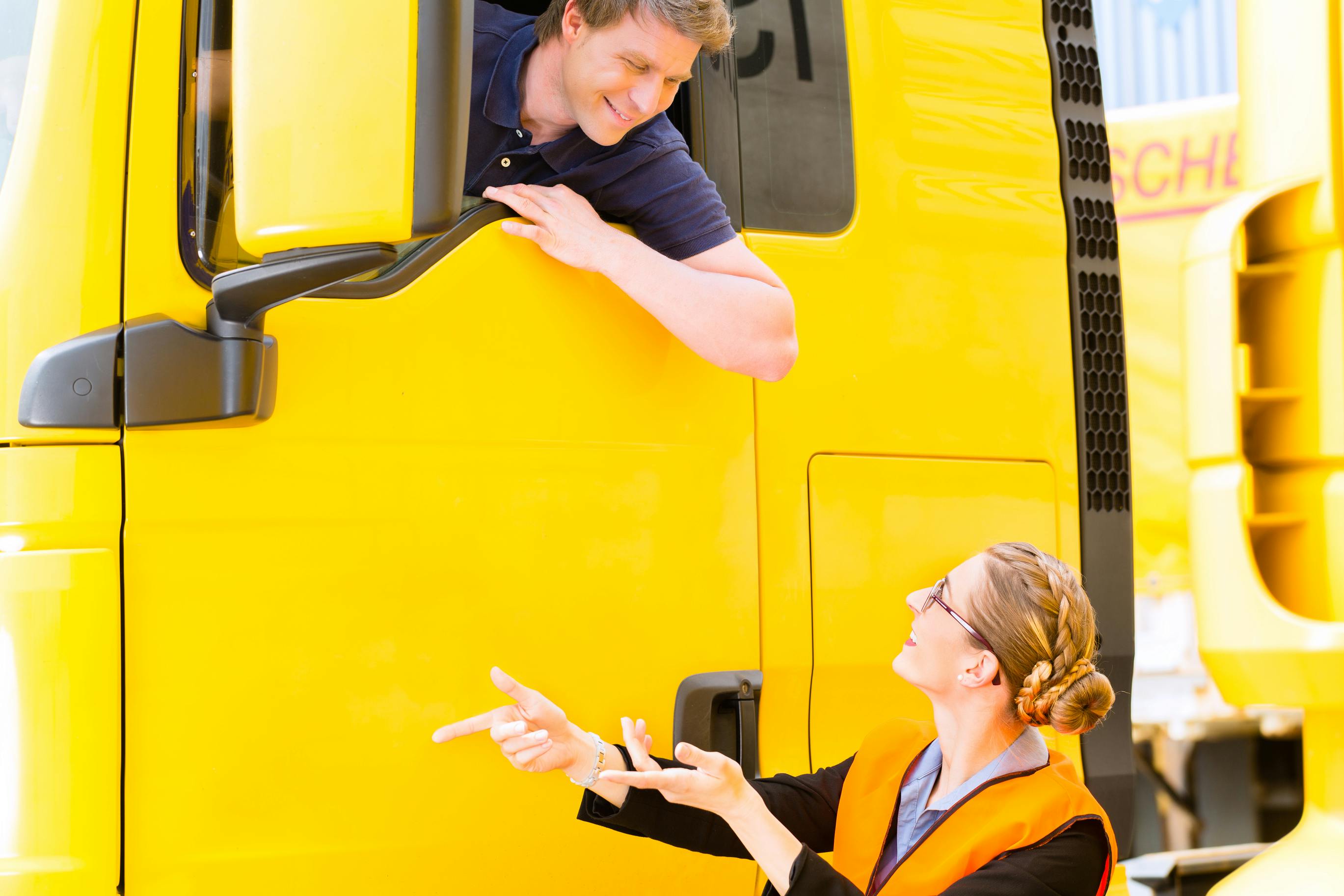 male truck driver in the cab talking to a woman wearing a safety vest on the ground