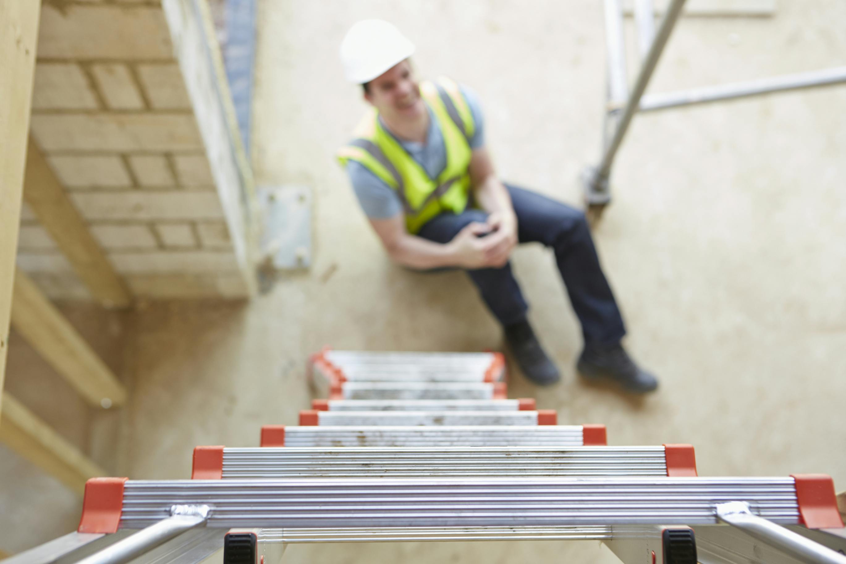 image of ladder in foreground with a man in hard hat and hi-vis safety vest clutching his knee on the ground
