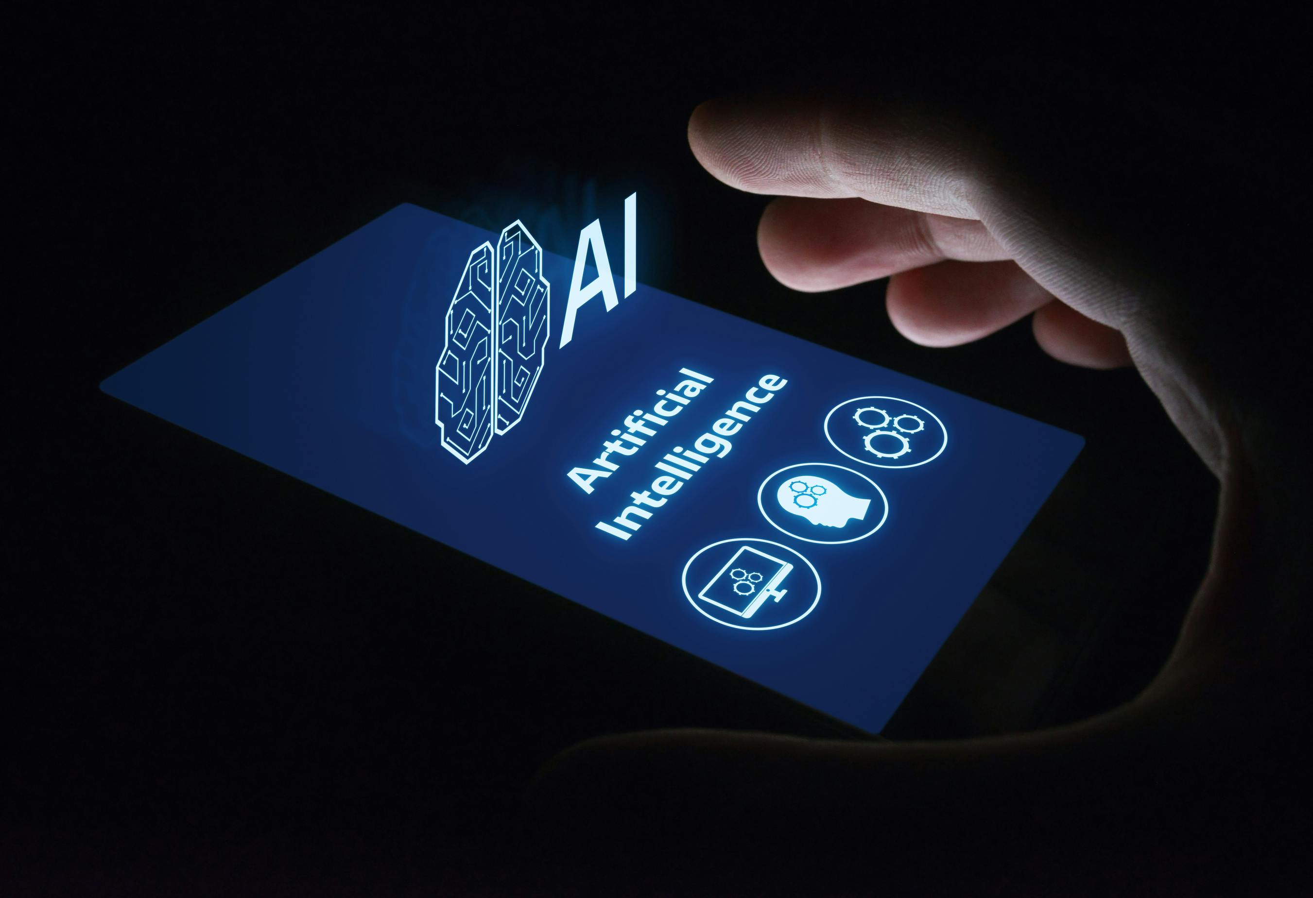 hand grasping smartphone that has a projection of the words artificial intelligence (AI) and symbols of a human head, computer screen and gears