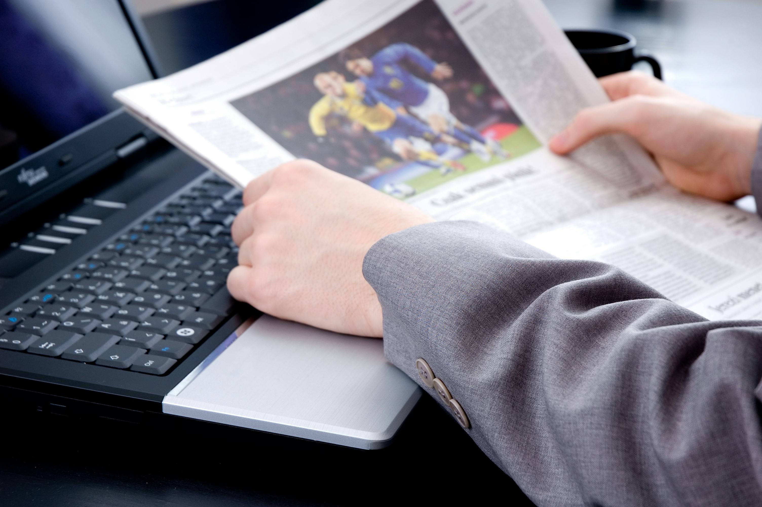 Image of someone in a suit coat reading the newspaper next to a laptop and coffee mug