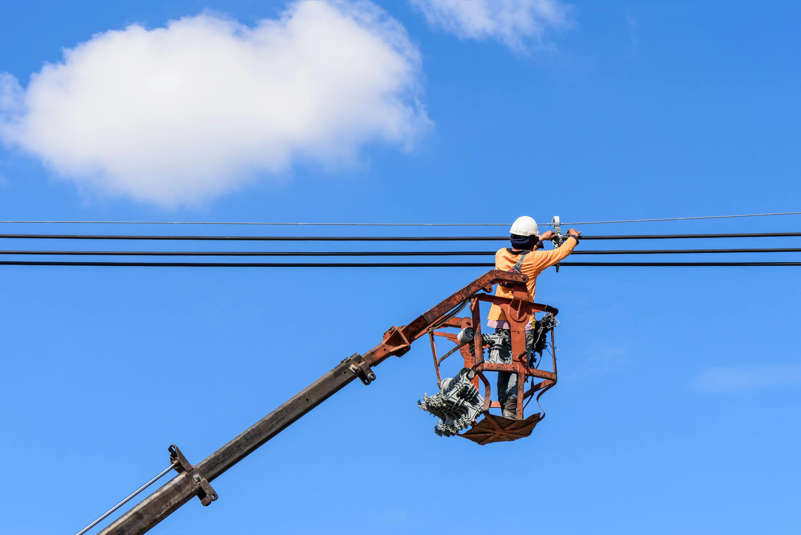 Field technician at height in bucket of hydraulic lift repairing electrical lines against a partly cloudy blue sky