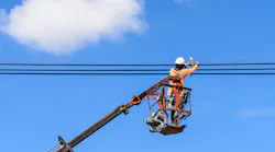 Field technician at height in bucket of hydraulic lift repairing electrical lines against a partly cloudy blue sky Field technician at height in bucket of hydraulic lift repairing electrical lines against a partly cloudy blue sky