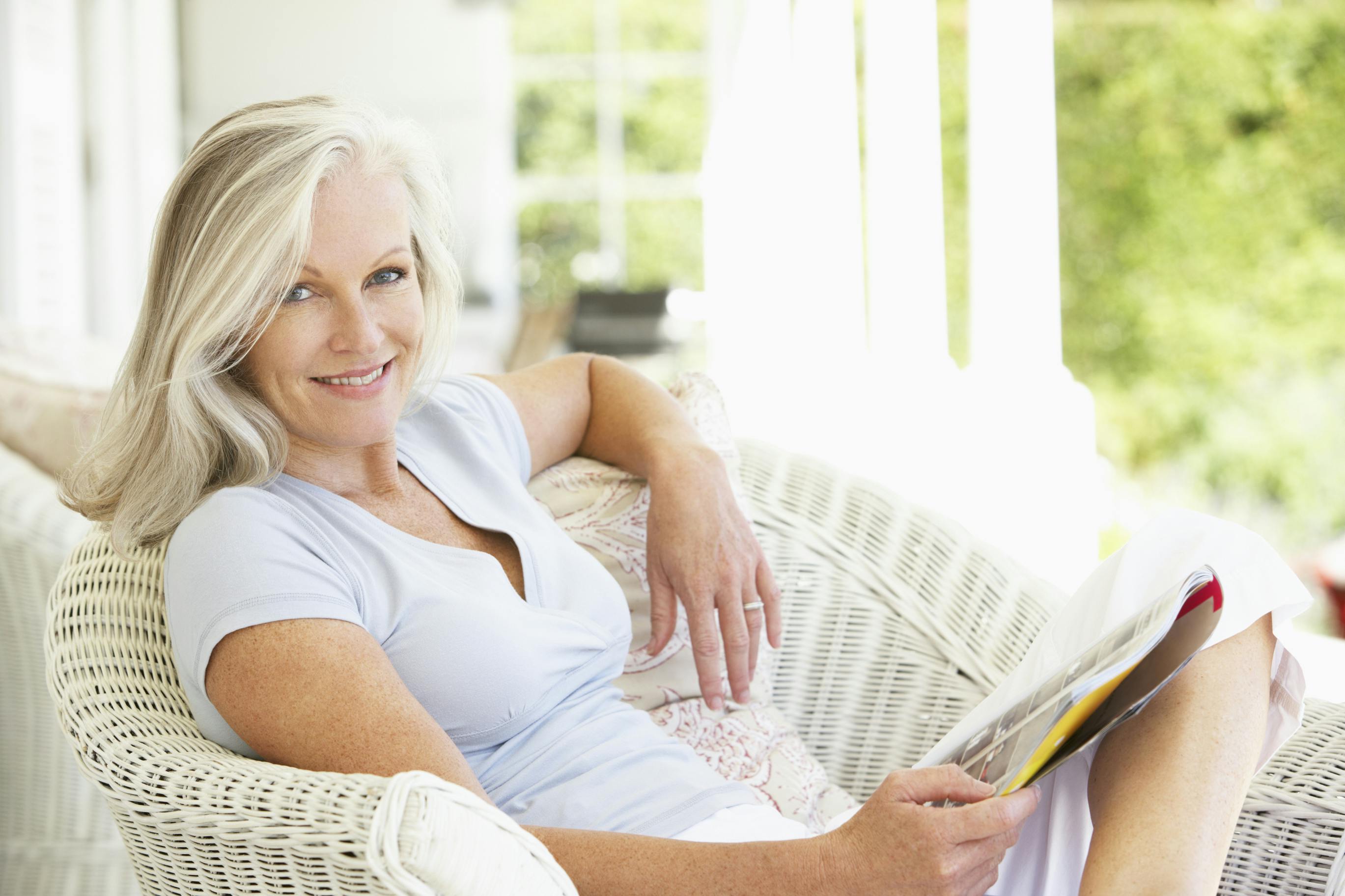 Woman with blonde hair sitting in a white wicker chair reading a magazine on a covered porch