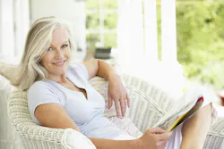 Woman with blonde hair sitting in a white wicker chair reading a magazine on a covered porch Woman with blonde hair sitting in a white wicker chair reading a magazine on a covered porch