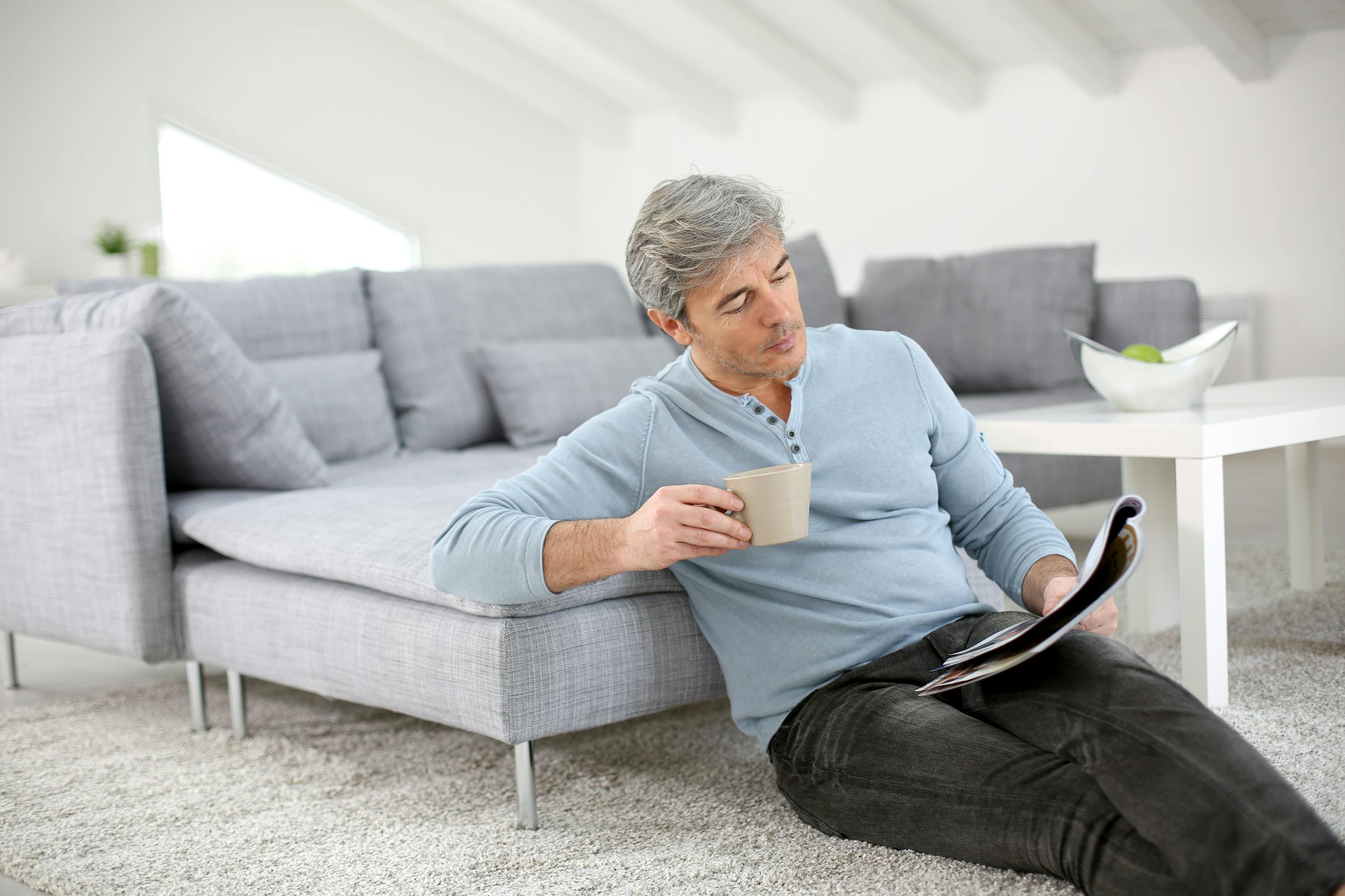 Image of man sitting on the floor with crossed legs holding a coffee cup, reading a magazine and leaning against a grey couch with chaise