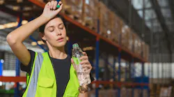 picture of woman wearing a hi-vis vest in a wearehouse wiping her forehead and drinking water, conveying she is hot