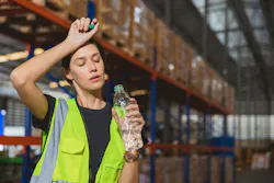 picture of woman wearing a hi-vis vest in a wearehouse wiping her forehead and drinking water, conveying she is hot