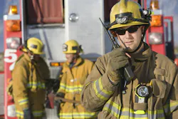 picture of a male firefighter on a walkie talkie with two male firefighters in the background next to fire truck