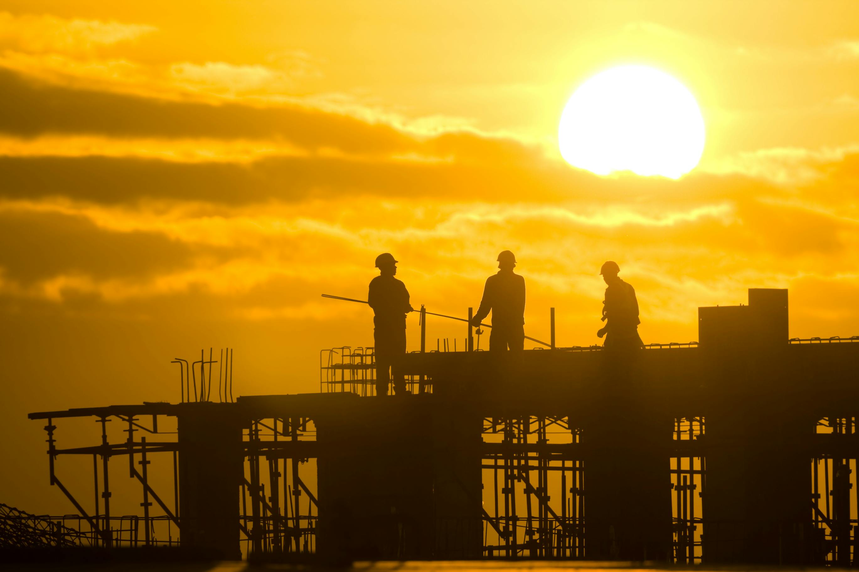 picture of three construction workers atop a framed structure with the sun in the background