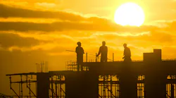 picture of three construction workers atop a framed structure with the sun in the background