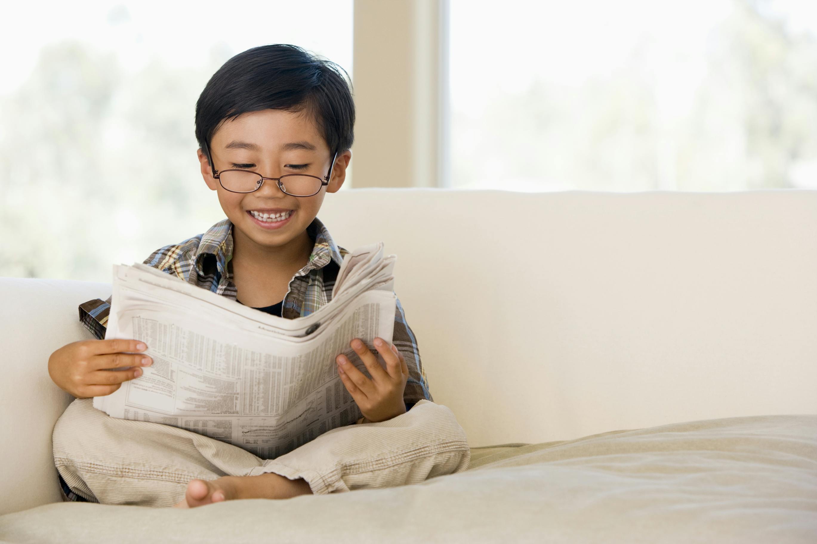 picture of a young boy with glasses sitting on a beige couch reading a newspaper