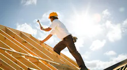 picture of man swinging a hammer at a wooden roof frame while working at height with sunshine and clouds behind him