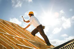 picture of man swinging a hammer at a wooden roof frame while working at height with sunshine and clouds behind him