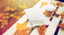 picture of a newspaper and to go coffee cup on a white bench strewn with vibrant fallen leaves