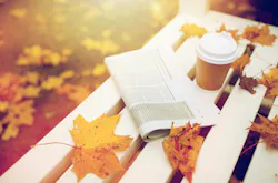 picture of a newspaper and to go coffee cup on a white bench strewn with vibrant fallen leaves